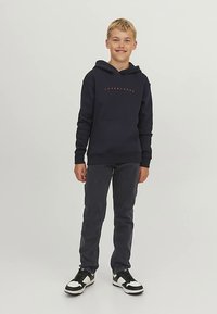 Sudadera con capucha azul marino con un bolsillo frontal y texto en naranja; combinada con jeans gris oscuro y zapatillas blancas y negras. Diseño simple y casual.