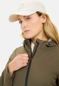Olive green jacket with a zippered front, adjustable hood, and elastic cuffs. Paired with a light beige cap featuring a logo.