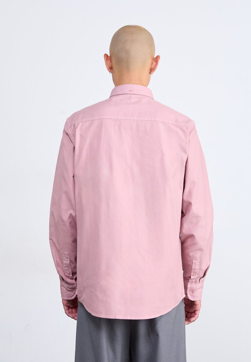 Camicia a maniche lunghe rosa chiaro con colletto abbottonato, caratterizzata da un orlo dritto e polsini abbottonati, indossata con pantaloni grigi.