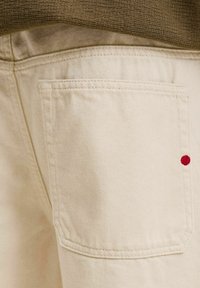 Gros plan de la poche arrière d’un pantalon beige avec un petit point rouge brodé près du bord droit et une partie supérieure marron texturée au-dessus.