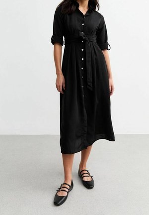 Femme portant une robe midi noire boutonnée avec manches retroussées et ceinture nouée à la taille, associée à des chaussures plates Mary Jane noires.