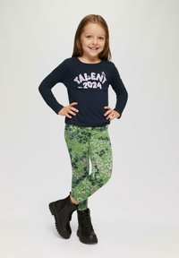 Marineblauwe longsleeve shirt met de opdruk "TALENT 2024", gecombineerd met groene bloemenleggings en zwarte enkellaarsjes met textuurzolen.