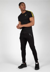 Zwart T-shirt met korte mouwen met gele streepdetail en logo; gecombineerd met zwarte tapse joggers met ritszakken en branding. Zwarte sneakers.