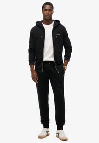 Felpa nera con zip e cordini sopra una maglietta bianca, abbinata a pantaloni jogger neri. Presenta dettagli con logo e sneakers bianche.