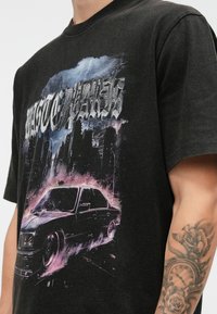 Crna majica s grafikom tamnog vintage automobila, gradskim obzorjem, munjom i stiliziranim tekstom, koju nosi osoba s tetoviranim podlakticama.