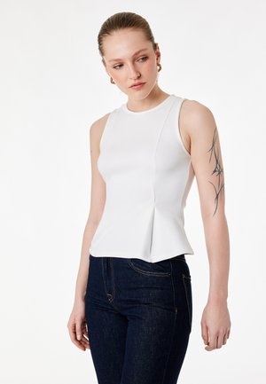 Mujer con el cabello recogido, vistiendo un top blanco sin mangas y jeans oscuros, mostrando un tatuaje abstracto negro en la parte superior de su brazo izquierdo.