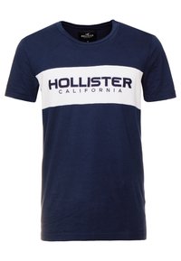 Mörkblå bomullströja med ett vitt horisontellt rand och "HOLLISTER CALIFORNIA" i mörkblå bokstäver över bröstet. Korta ärmar.