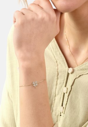 Gold chain bracelet featuring a four-leaf clover design set with clear gemstones, worn on a wrist with a light green buttoned blouse.