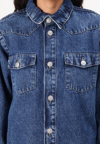 Cămașă din denim cu un guler clasic, două buzunare pe piept cu închidere cu nasturi și accesorii argintii. Țesătura are o textură bleu decolorată.