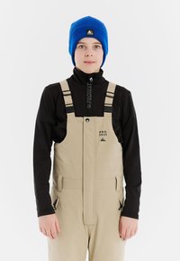 Kind in beige bretels en een zwart shirt met lange mouwen, met een blauwe beanie, staande tegen een witte achtergrond.
