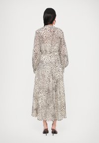 Hosszú, áttetsző ruha bézs alapon fekete és barna pöttyös mintával, hosszú ujjakkal és derékrészen összeszűkülve, magassarkú szandállal párosítva.