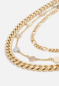 Collana multi-strato in tonalità dorata con catene spesse e sottili, accenti esagonali e circolari e decorazioni brillanti su alcuni collegamenti.