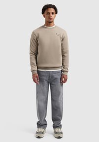 Beige sweatshirt met ronde hals en reliëflogo aan de linkerkant. Gecombineerd met lichtgrijze jeans en sportieve beige en zwarte sneakers.