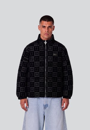 Mann trägt eine schwarze, gemusterte Jacke mit Reißverschluss und hohem Kragen sowie hellblaue, lockere Jeans und steht vor einem einfarbigen grauen Hintergrund.