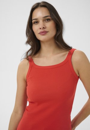 Jeune femme aux longs cheveux bruns portant un débardeur rouge sans manches sur un fond clair uni.