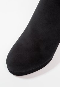 Gros plan sur le bout arrondi et la partie supérieure d'une botte en suède noir sur un fond blanc.