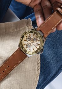 Auksinė ir sidabrinė aštuntakampė laikrodžio dėžutė su ruda odine apyranke, trimis subdizainais ir kreminiu ciferblatu, rodantis valandų žymeklius ir prekės pavadinimą.