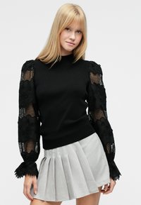 Maglione nero con maniche a rete testurizzate con motivi floreali. Gonna grigia plissettata completa l'outfit.