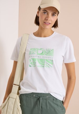 Frau trägt ein weißes "Coastal Garden" T-Shirt, khakifarbene Kordelzughose, eine beige Kappe und trägt eine beige Umhängetasche.