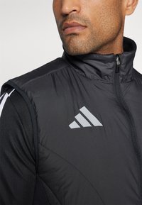 Chaleco sin mangas negro con un cuello alto, hecho de tejido acolchado. Presenta un logo gris de Adidas en el pecho y detalles en tela negra en contraste.