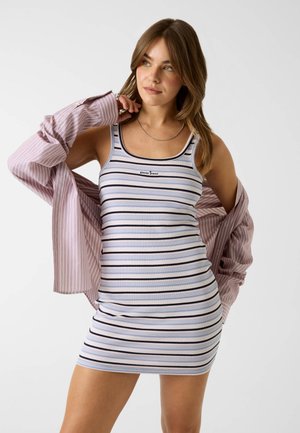 Femme portant une mini-robe sans manches rayée avec un "guess jeans" et une chemise ample rayée rose, regardant sur le côté avec la main près du cou.