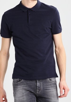 Mann trägt ein eng geschnittenes dunkelblaues Polohemd mit kurzen Ärmeln und hellgraue verwaschene Jeans, steht vor einem schlichten Hintergrund.