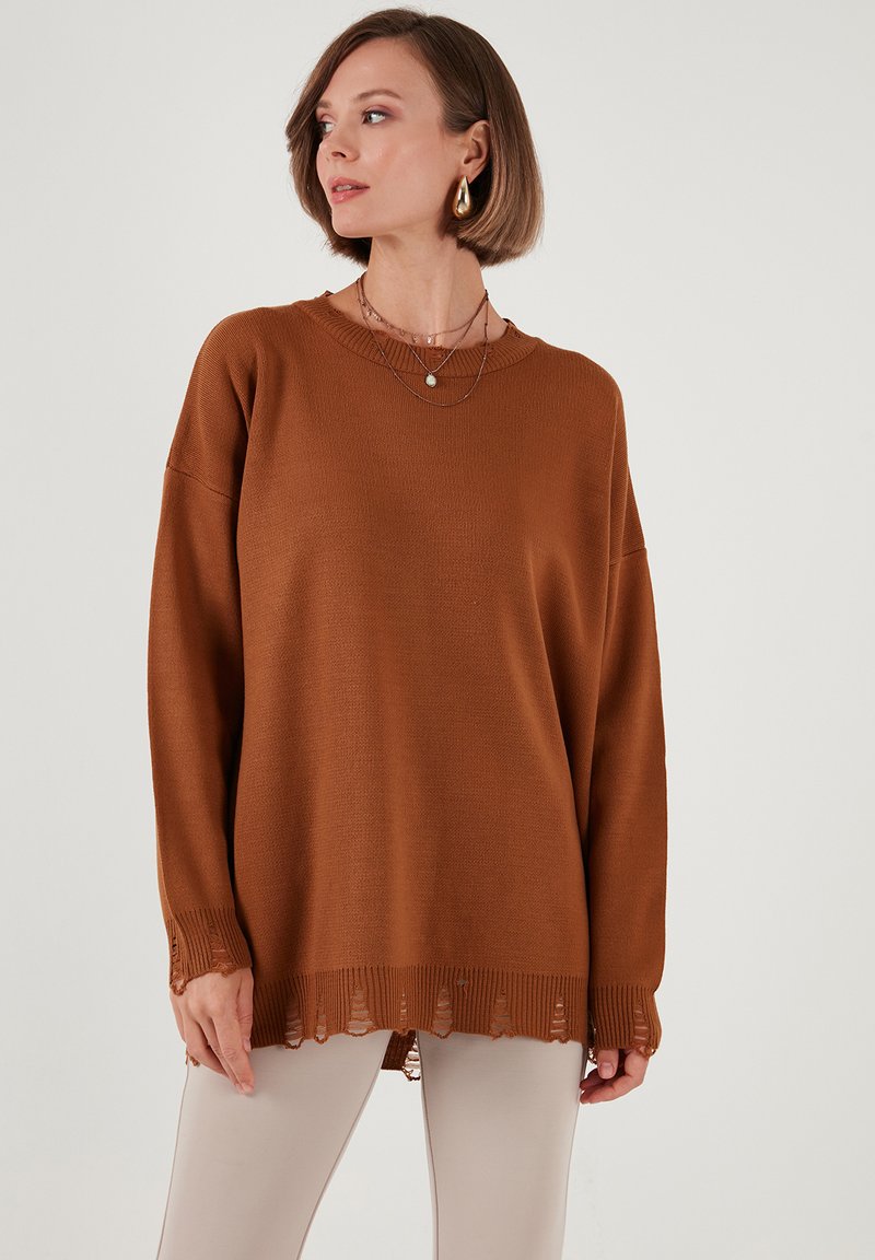 Maglione marrone oversize con texture a coste, spalle cadenti e orlo scallop. Abbinato a leggings chiari e collane a strati.
