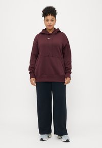 Hoodie oversized em borgonha com bolso frontal, capuz com cordão e logótipo branco; combinado com calças largas azuis de cintura alta e ténis claros.