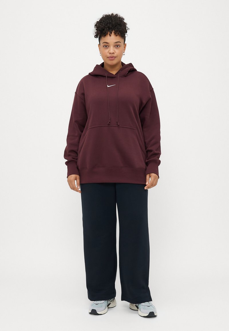 Burgunderfarbenes übergroßes Hoodie mit einer Fronttasche, Kapuze mit Kordelzug und weißem Logo; kombiniert mit navyblauen weit geschnittenen Hosen und hellen Sneakern.