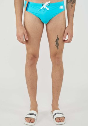 Man draagt felblauwe zwembroek met zwarte en rode strepen, witte trekkoord, beentattoo en witte slides met zwart logo.