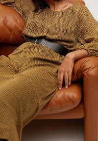 Długa, brązowa sukienka w kropki z lekkiego materiału, przewiązana szerokim czarnym paskiem. Osoba odpoczywa na brązowej skórzanej sofie.