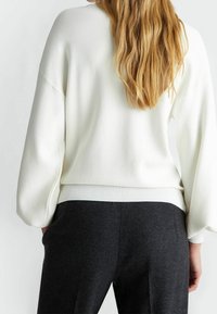 Femme aux longs cheveux blonds portant un pull blanc à manches longues et un pantalon gris foncé, se tenant avec les mains dans les poches, tournant le dos.