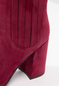 Dieprode suède enkellaars met verticale plooien, een vierkante teen en een chunky hak. Heeft contrasterende stiksel en een gladde textuur.