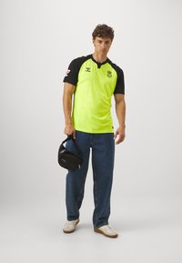 Maglietta da calcio giallo neon con accenti neri, maniche corte e colletto. Jeans blu scuro larghi e una borsa nera tenuta nella mano destra.