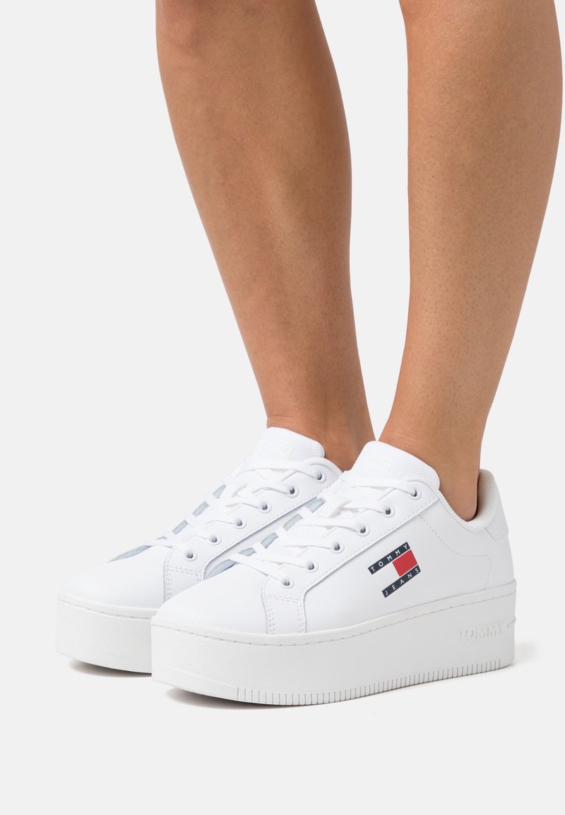 Jambes portant des baskets blanches à plateforme avec lacets, avec un logo Tommy Jeans sur le côté et une semelle épaisse.