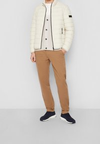 Giacca puffer bianco sporco con colletto alto e zip nera, abbinata a una camicia grigia chiara con bottoni e pantaloni chino beige. Sneakers scure.