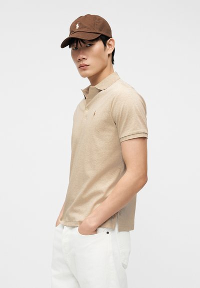 Νεαρός άνδρας που φοράει μπεζ πουκάμισο πόλο και καφέ καπέλο, στέκεται με τα χέρια στις τσέπες λευκών παντελονιών, μπροστά σε απλό φόντο.
