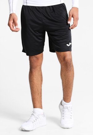 Mann trägt schwarze Sportshorts mit einem weißen Logo am linken Bein, weiße Turnschuhe und weiße Socken, steht vor einem schlichten Hintergrund.