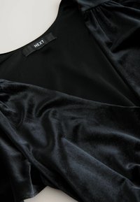 Black velvet garment with a smooth texture, featuring a deep neckline and soft sleeves. Label reads "NEXT." The fabric shows subtle light reflections.