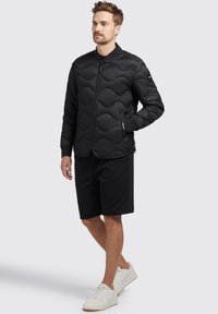 Black quilted jacket with wave pattern, front zipper, and knit cuffs, paired with black shorts and white sneakers. Simple and functional design.