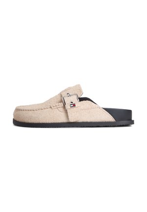 Mule beige texturizado tipo slip-on con suela negra y hebilla lateral que presenta un pequeño detalle de logo en rojo y azul.