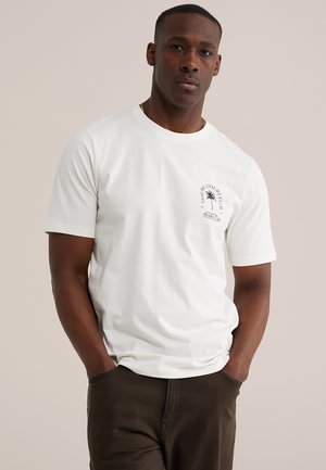 Muž s tmavou pokožkou v bielom tričku s malým čiernym logom palmy a textom na hrudi, ruky v tmavých vreckách nohavíc.