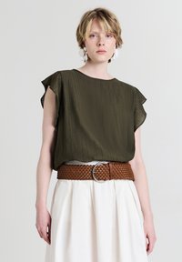 Giovane con capelli biondi corti che indossa una maglietta verde oliva con maniche a farfalla, una larga cintura di tessuto marrone, una gonna piegata bianca e orecchini floreali.