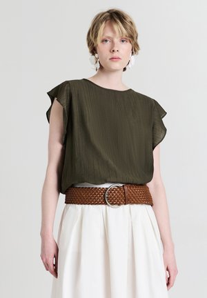 Giovane con capelli biondi corti che indossa una maglietta verde oliva con maniche a farfalla, una larga cintura di tessuto marrone, una gonna piegata bianca e orecchini floreali.