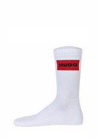 Weißer Baumwollsocken mit geripptem Muster. Verfügt über ein rotes rechteckiges Patch mit "HUGO" in schwarzer Schrift. Design bis zur Mitte der Waden.