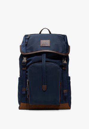 Mochila azul marino con bolsillo frontal con cremallera, borde inferior marrón, solapa superior con cierre, bolsillos laterales y parche con el logo de Aldo en la solapa.