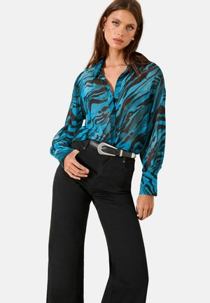 Žena nosi dlhú blúzku s modrými a čiernymi tigrovými pruhmi, zastrčenú do čiernych nohavíc s vysokým pásom a širokým opaskom so striebornou prackou.