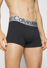 Crne pamučne bokserice s srebrnim Calvin Klein logotipom na pojasu. Imaju prilagođen fit i glatku teksturu.