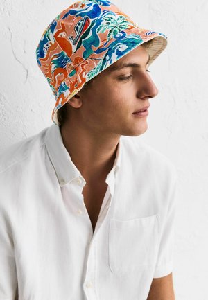 Young man wearing a white button-up shirt and a colorful bucket hat with abstract orange, blue, and green patterns, facing right against a white wall.