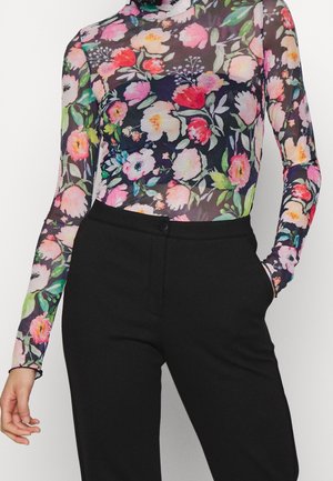 Mujer con una camiseta ajustada de manga larga transparente con flores en tonos rosas, rojos y verdes, combinada con pantalones negros de talle alto y corte a medida.